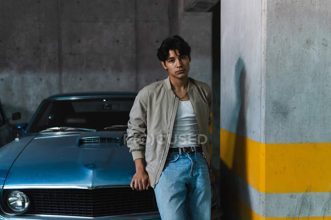 Portrait de jeune homme latin en vêtements décontractés regardant avec confiance la caméra tout en s'appuyant sur une voiture vintage dans un parking — Photo de stock