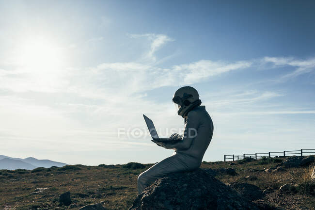 Full body male astronaut in spacesuit browsing data on netbook while sitting on rock outside station with rocket shaped antennas — Stock Photo