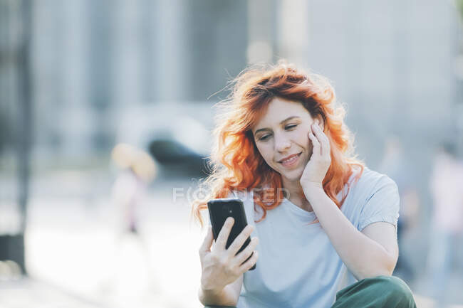 Vista lateral alegre pelirroja mujer sentada en la calle y mensajería en las redes sociales en el teléfono móvil - foto de stock