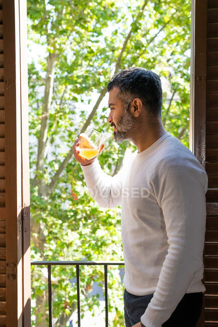 Мужественный бородатый мужчина, пьющий апельсиновый сок из стекла на балконе в солнечный день — стоковое фото