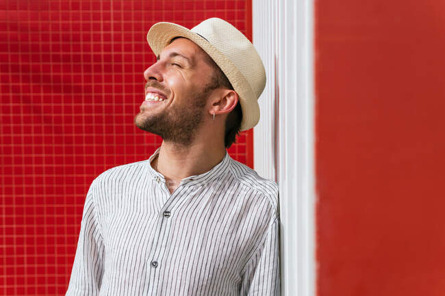 Счастливый молодой бородатый мужчина в стильной полосатой рубашке и соломенной шляпе ярко улыбается, стоя возле красной стены и наслаждаясь солнечным летним днем на улице — стоковое фото