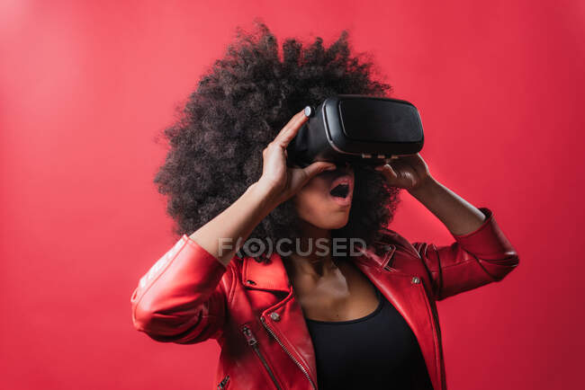 Astonished African American female with opened mouth and in VR goggles experiencing virtual reality on red background in studio — Stock Photo