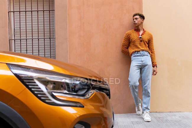 Joven chico de pelo rizado étnico con estilo en traje de moda apoyado contra la pared cerca de aparcado moderno automóvil naranja en la calle urbana - foto de stock