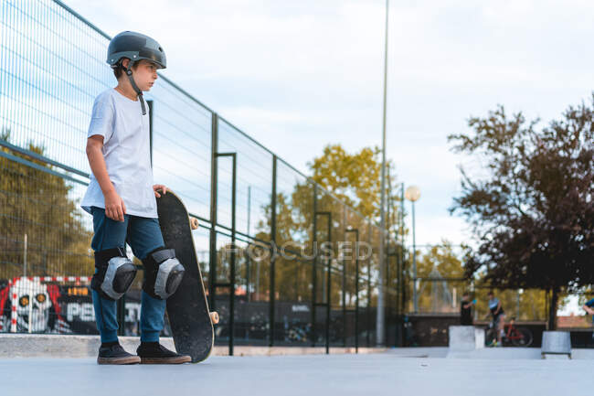 Вид збоку впевненого хлопчика-підлітка зі скейтбордом і в захисному шоломі, що стоїть в скейт-парку і дивиться в сторону — стокове фото