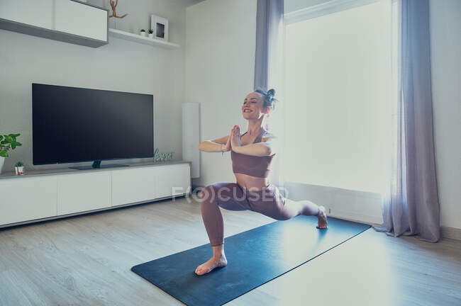 Young content female standing with namaste hands on yoga mat while looking forward in house room in sunlight — Stock Photo