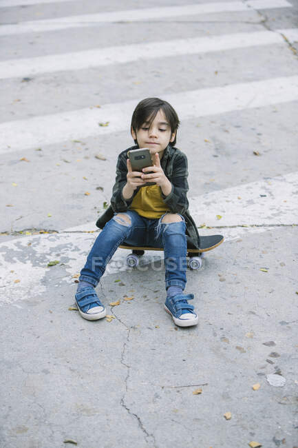 Зверху повне тіло чарівного хлопчика в повсякденному одязі переглядаючи мобільний телефон, сидячи на скейтборді на пішохідному переході в місті — стокове фото