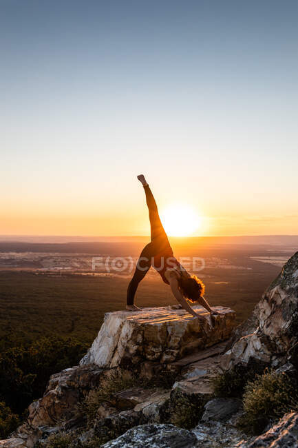 Joven yogi practicando yoga sobre una roca en la montaña con la luz del amanecer, vista lateral - foto de stock