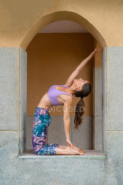 Vue latérale de la jeune femme pieds nus en vêtements de sport montrant la pose d'Ustrasana tout en pratiquant le yoga et en levant les yeux entre les trous voûtés — Photo de stock
