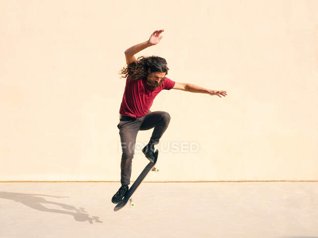 Männlicher Skateboarder mit welligem Haar führt Trick auf Skateboard aus, während er über Gehweg springt und an sonnigen Tagen nach unten schaut — Stockfoto