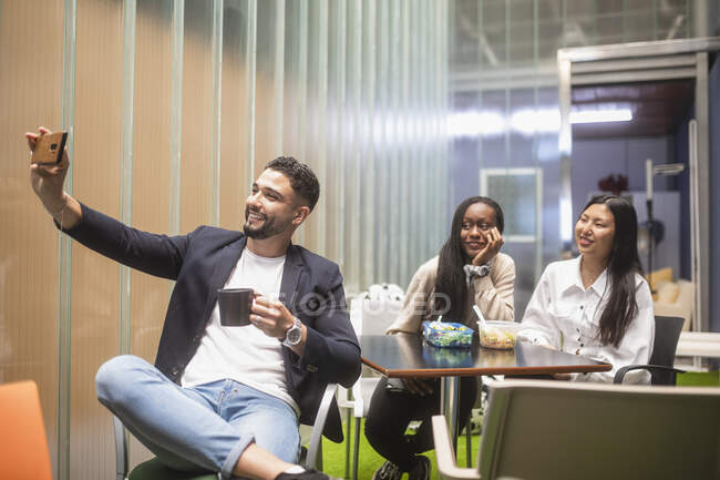 Gruppe fröhlicher, multiethnischer Kollegen, die sich während des Mittagessens am Tisch im Arbeitsbereich mit dem Smartphone fotografieren — Stockfoto