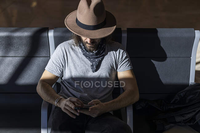 The guy in the hat at the airport in the waiting room sitting waiting for his flight, with wireless headphones to listen to music while chatting with his smart phone, top view — Stock Photo