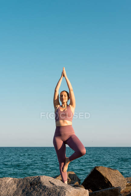 Donna calma in piedi in Vrksasana con gli occhi chiusi e praticare yoga sulla pietra sulla spiaggia vicino al mare — Foto stock