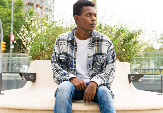 Афроамериканский стильный мужчина сидит на скамейке с розетками для зарядки устройств в городе и смотрит в сторону — стоковое фото