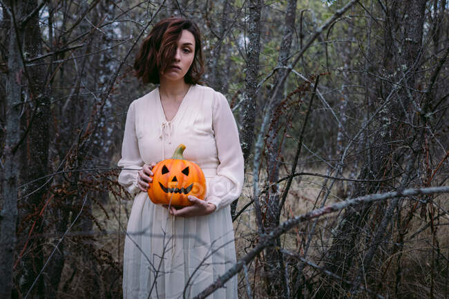 Female in white dress standing with glowing pumpkin lantern in the woods on Halloween and looking at camera — Stock Photo
