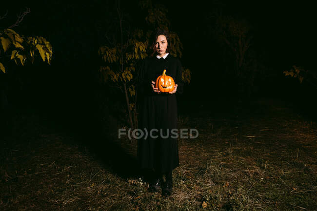 Donna in abito nero in piedi con lanterna di zucca incandescente in boschi scuri su Halloween e guardando la fotocamera — Foto stock