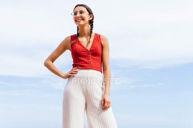 Baixo ângulo de fêmea alegre na roupa de verão na moda em pé com a mão na cintura no fundo do céu nublado azul — Fotografia de Stock
