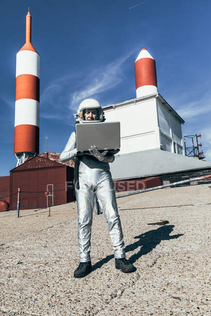 Astronauta macho de cuerpo completo en traje espacial navegando datos en netbook mientras está de pie fuera de la estación con antenas en forma de cohete - foto de stock