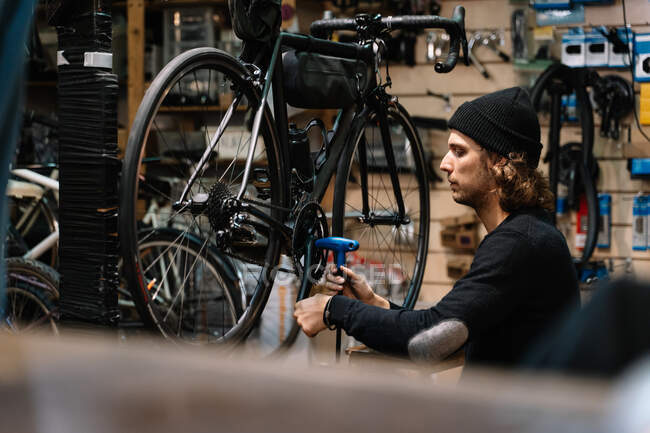 Vista lateral de un joven mecánico masculino experto con rueda de cadena de montaje de herramientas en bicicleta durante los trabajos de servicio de reparación en el taller - foto de stock
