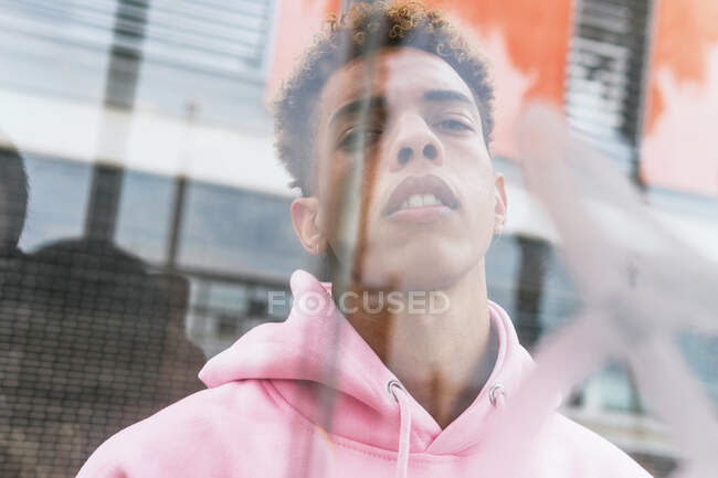 Através de vidro de sério jovem etnia hipster cara com penteado afro vestido com capuz rosa na janela e olhando para a câmera — Fotografia de Stock