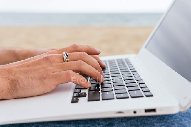 Cropped unrecognizable male blogger in stylish wear and hat sitting on sandy beach and typing on laptop during summer holidays on seashore — Stock Photo