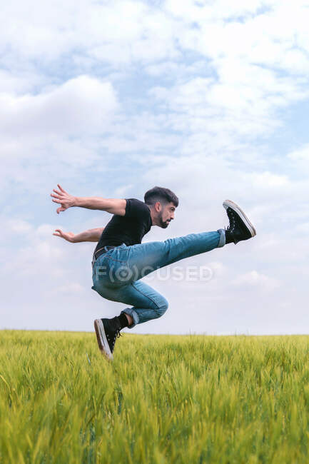 Hombre vista lateral en el salto de mezclilla con la pierna levantada por encima de la hierba alta en el campo sombrío - foto de stock