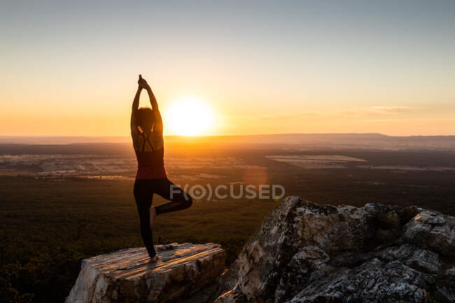 Giovane donna yogi che pratica yoga su una roccia in montagna con la luce dell'alba, vista posteriore con una gamba e le braccia sollevate — Foto stock