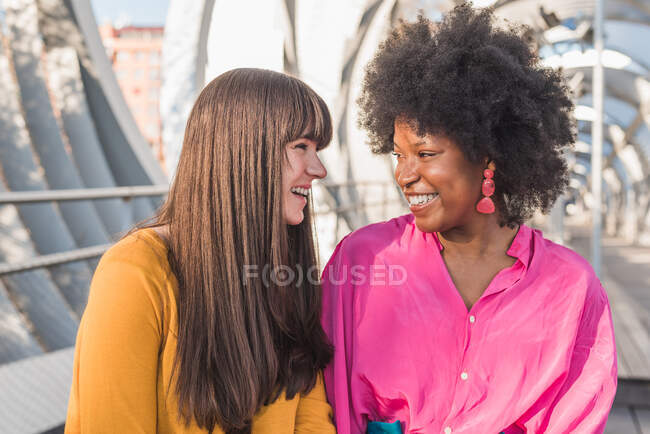 Щаслива багатоетнічна ЛГБТ пара жінок тримається за руки, стоячи на мосту в місті в сонячний день і дивлячись один на одного — стокове фото