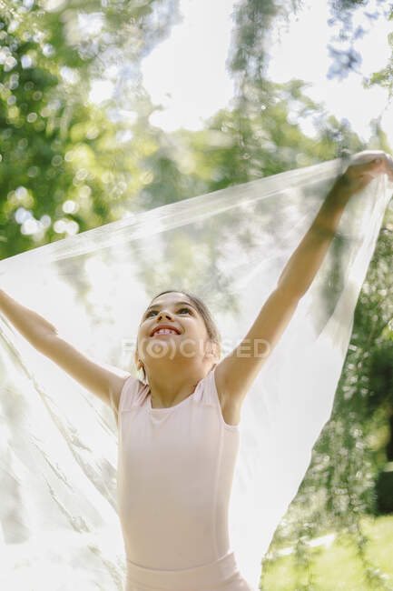 Зміст дівчини-підлітка в балетній сукні, що грає з прозорою тканиною на лузі в парку в сонячний день — стокове фото