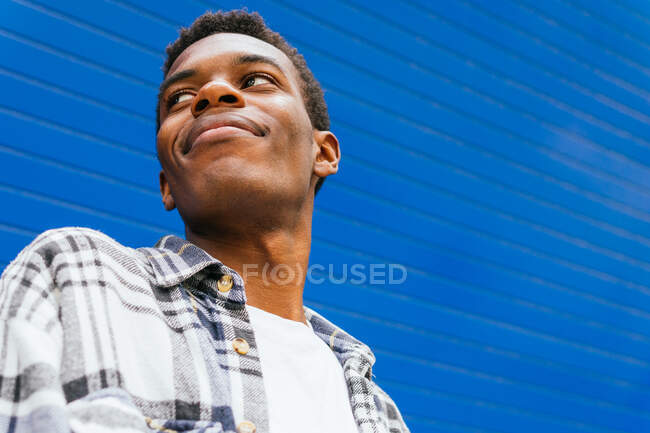 Low angle of handsome smiling African American male looking away on bright blue background in street in summer — Stock Photo