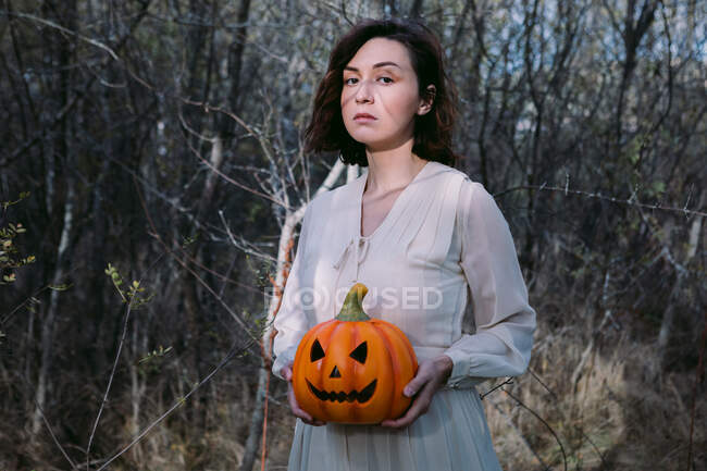 Female in white dress standing with glowing pumpkin lantern in the woods on Halloween and looking at camera — Stock Photo