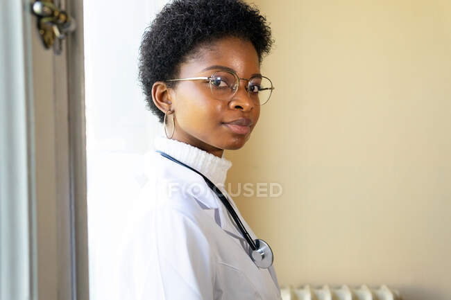 Побочный обзор компетентной молодой афроамериканской женщины-врача в белом медицинском халате и очках со стетоскопом, смотрящей в камеру, стоя в клинике — стоковое фото