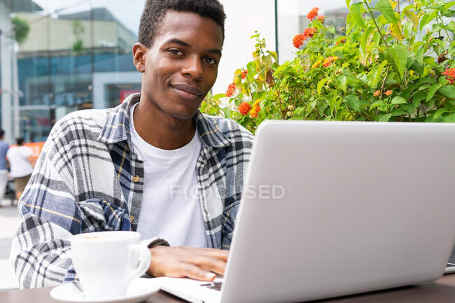 Feliz afroamericano freelancer masculino navegando y trabajando remotamente en el ordenador portátil en la cafetería al aire libre mientras está sentado mirando a la cámara en la mesa con una taza de café - foto de stock