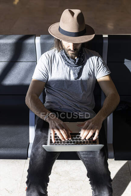 El tipo en el sombrero en el aeropuerto en la sala de espera sentado esperando su vuelo, con auriculares inalámbricos para escuchar música mientras trabaja con su computadora portátil, vista superior - foto de stock