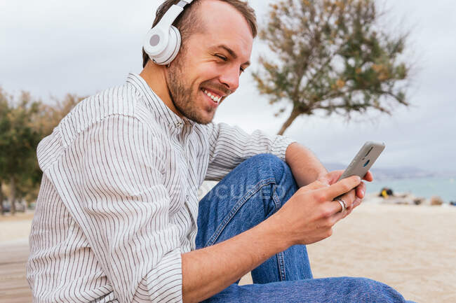 Vista lateral do jovem macho barbudo alegre em fones de ouvido sem fio ouvindo música e navegando telefone celular enquanto descansa sozinho na praia de areia no dia de verão — Fotografia de Stock