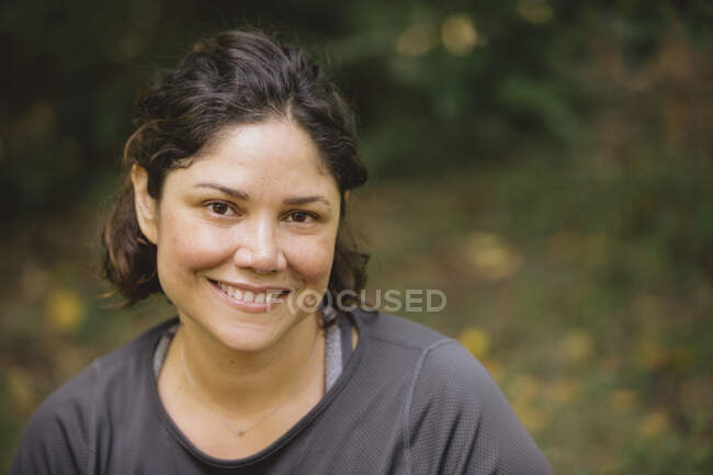 Позитивна молода етнічна жінка з коричневим кучерявим волоссям в повсякденному одязі посміхається і дивиться на камеру, відпочиваючи в зеленому парку під час вихідних — стокове фото