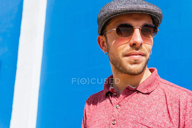 Молодий бородатий хлопець в повсякденній червоній сорочці поло і кепці стоїть на синій стіні на вулиці в літній день — стокове фото