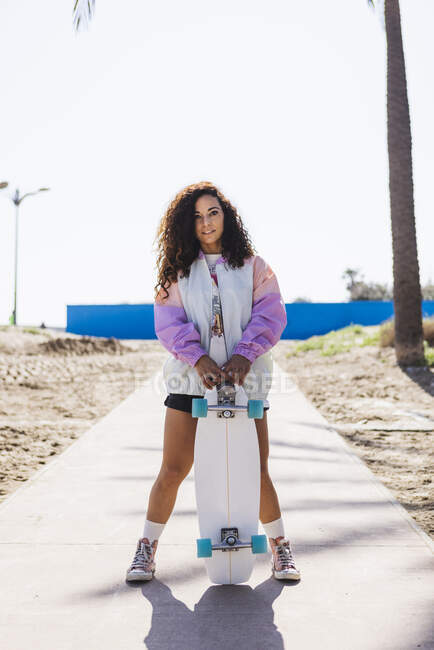 Повне тіло жінки зі скейтбордом в руці стоїть, дивлячись на камеру на тротуарі уздовж високих долонь на узбережжі і морі — стокове фото