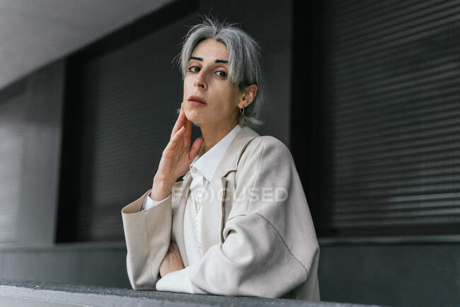 Serious transgender female in trendy coat leaning on gray fence while standing in street looking at camera — Stock Photo