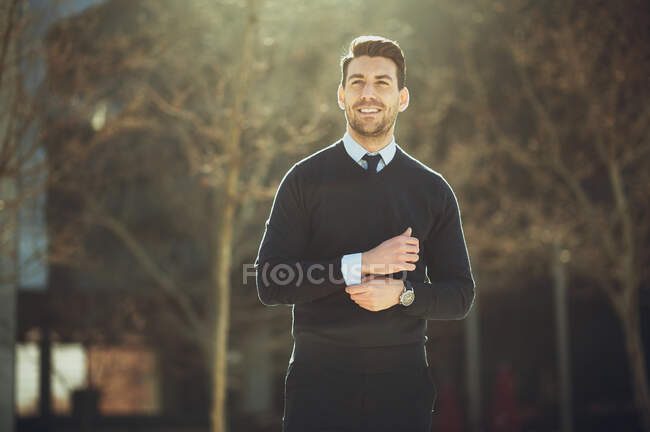 Улыбающийся бородатый мужчина-предприниматель в наручных часах с современной стрижкой, смотрящий в город в подсветке — стоковое фото