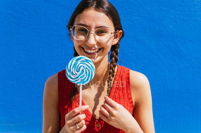 Positive charming female standing with sweet lollipop in street on sunny day on blue background and looking at camera — Stock Photo
