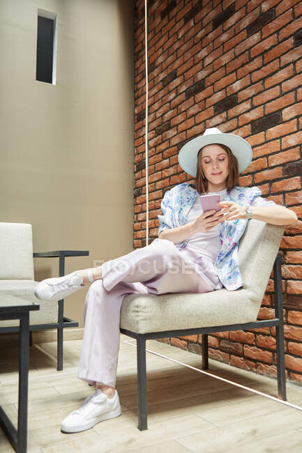 Trendy young female in hat text messaging on cellphone while sitting with crossed legs on soft chair in building — Stock Photo