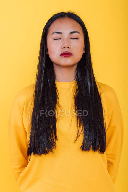 Retrato de mujer asiática de pie sobre fondo amarillo en estudio con los ojos cerrados - foto de stock