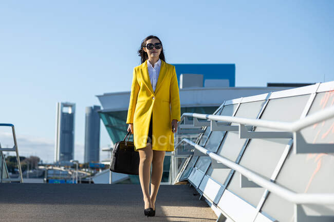Femme d'affaires asiatique avec manteau jaune marchant dans la rue avec bâtiment en arrière-plan — Photo de stock