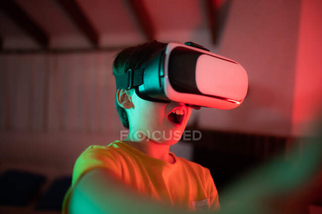 Unrecognizable concentrated surprised little boy experiencing virtual reality in modern headset in dark room — Stock Photo