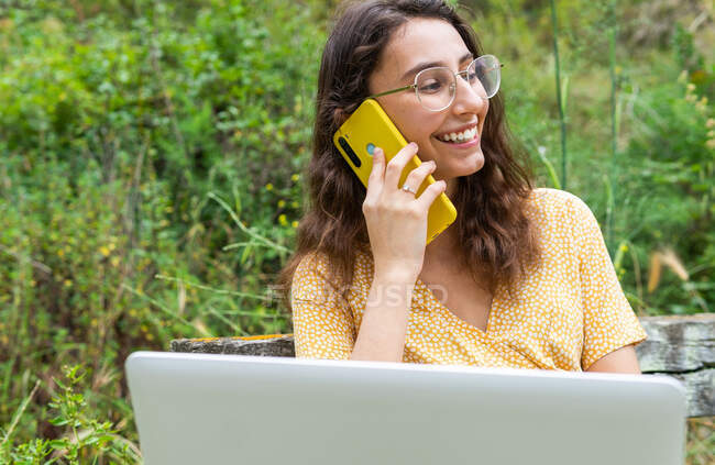 Delighted female freelancer having mobile phone conversation while sitting with laptop in summer park and discussing project — Stock Photo