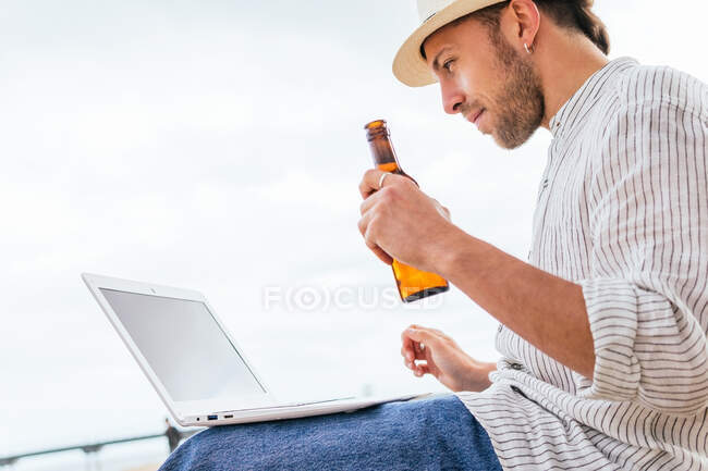 Снизу вид на молодого блогера в стильной одежде и шляпе, сидящего с бутылкой пива на песчаном пляже и печатающего на ноутбуке во время летних каникул на берегу моря — стоковое фото
