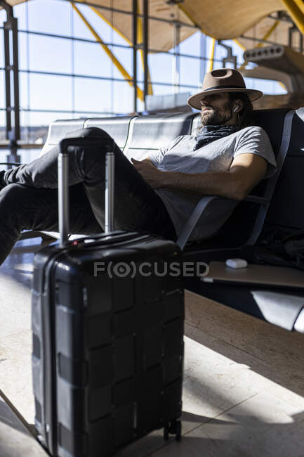 Le gars avec un chapeau à l'aéroport dans la salle d'attente assis en attendant son vol, avec des écouteurs sans fil pour écouter de la musique, dort et avec son chapeau couvre ses yeux — Photo de stock
