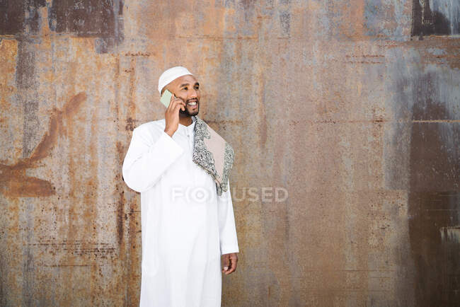 Веселый мусульманский мужчина в традиционной одежде улыбается и разговаривает по мобильному телефону, стоя возле потрепанной стены на улице — стоковое фото