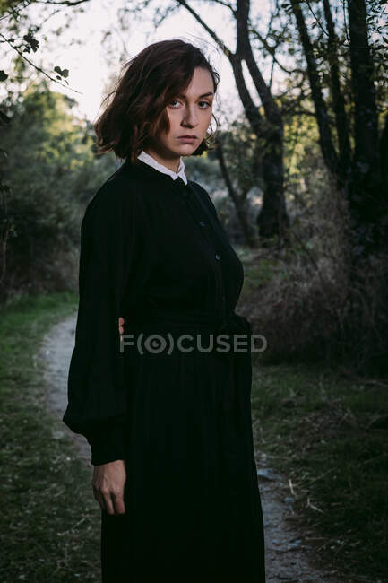 Mulher sem emoção vestindo vestido preto longo em pé na floresta de outono durante a celebração do Halloween olhando para a câmera — Fotografia de Stock