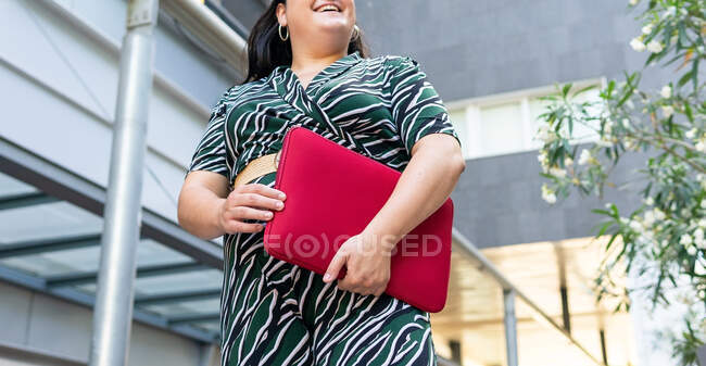From below anonymous cheerful young female in stylish striped outfit with red laptop case in hands while standing against contemporary city building — Stock Photo